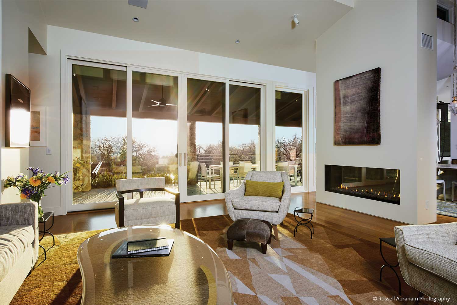 A sunset shines through glass sliding doors into a furnished sitting room. Artwork is displayed above a nearby fireplace.