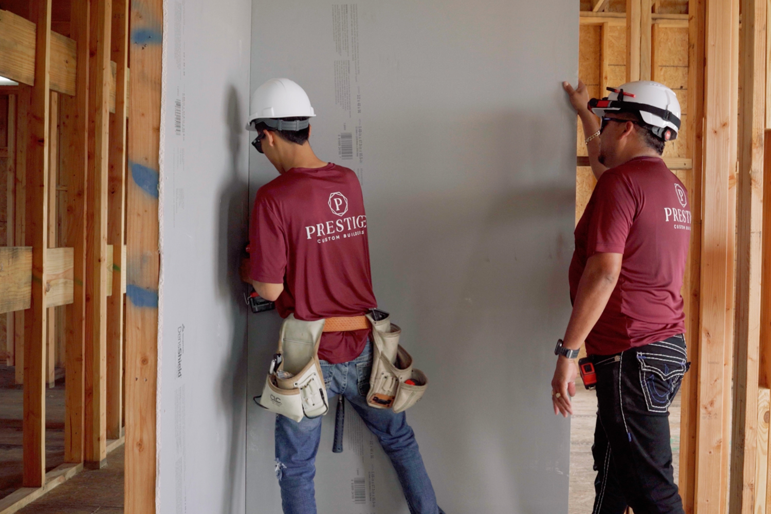 Two members of Prestige Custom Builders wearing hard hats and maroon t-shirts install DensShield® Tile Backer in a home. 