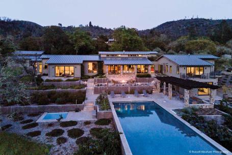 A residence emits warm light through its windows and doors. It is surrounded by views of desert foliage and a pool.